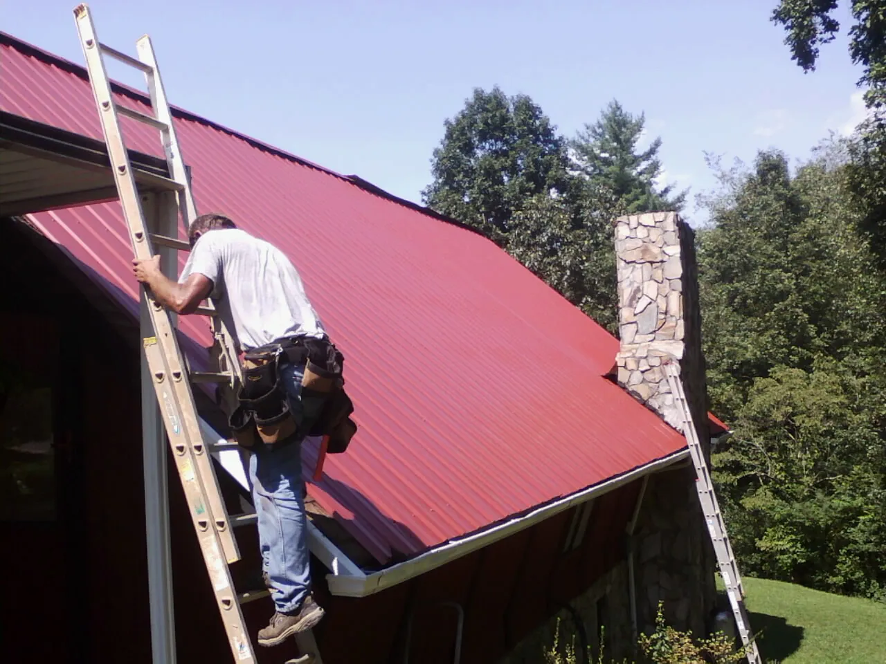 Roofer on a ladder beside a red metal roof and stone chimney — Soffit & Fascia Repair by Roofing Brothers in Saint Helena Island